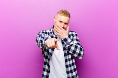 Kare gömlekli genç sarışın adam kamerayı göstererek sana gülüyor ve seninle dalga geçiyor ya da alay ediyor.