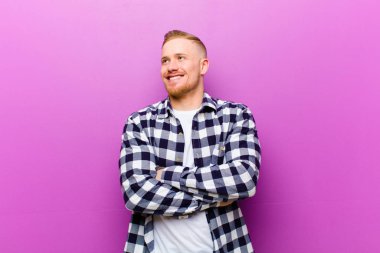 Kare gömlekli genç sarışın adam mutlu, gururlu ve umutlu hissediyor, merak ediyor ya da düşünüyor, çapraz kollarıyla fotokopi çekmeye bakıyor.