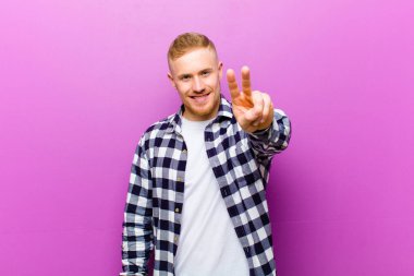 Kare gömlekli genç sarışın adam gülümsüyor ve mutlu görünüyor, kaygısız ve pozitif, zafer ya da barış işareti yapıyor.