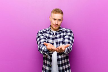 Kare gömlekli genç sarışın adam mutlu bir şekilde gülümsüyor arkadaş canlısı, kendinden emin, pozitif bakışlı, bir nesne ya da konsept sunuyor ve gösteriyor.