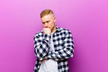 Kare gömlekli genç sarışın adam ciddi, düşünceli ve endişeli, elleri çenesine bastırılmış bir şekilde yan bakıyor.