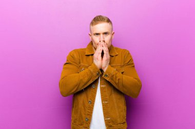 Genç sarışın adam mutlu ve heyecanlı, şaşırmış ve hayrete düşmüş elleriyle ağzını kapatmış, mor arka plana karşı sevimli bir ifadeyle kıkırıyor.