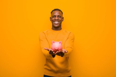 Genç Afrikalı Amerikalı siyah adam turuncu duvara karşı domuz kumbarası ile