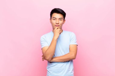 Genç Çinli adam mutlu ve gülümsüyor, çenesi üzerinde, merak ediyor ya da soru soruyor, düz renkli duvarla seçenekleri karşılaştırıyor.