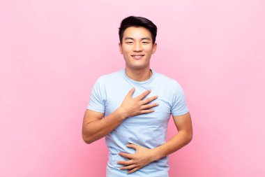 Genç Çinli bir adam komik bir şakaya yüksek sesle gülüyor, mutlu ve neşeli hissediyor, düz renkli duvara karşı eğleniyor.