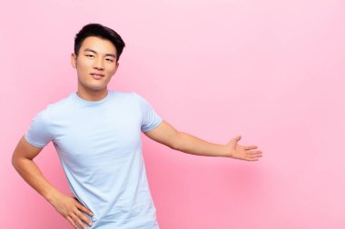 Genç Çinli adam kendini mutlu ve neşeli hissediyor, gülümsüyor ve seni selamlıyor, düz renkli duvara karşı dostça bir jestle içeri davet ediyor.