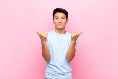 Genç Çinli adam neşeyle gülümsüyor ve mutlu görünüyor, kaygısız ve pozitif hissediyor iki başparmağı düz renkli duvarda