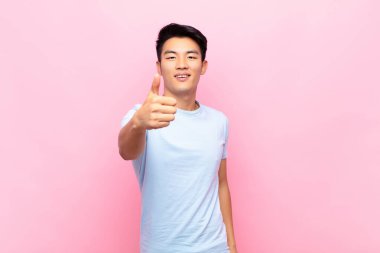 Genç Çinli adam gururlu, kaygısız, kendinden emin ve mutlu, parmakları düz renkli duvara değmiş gülümsüyor.