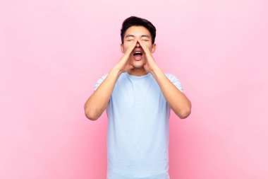 Genç Çinli adam mutlu, heyecanlı ve pozitif hissediyor. Elleri ağızda, düz renkli duvara karşı bağırıyor.