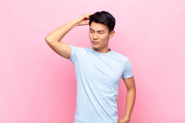 young chinese man feeling puzzled and confused, scratching head and looking to the side against flat color wall