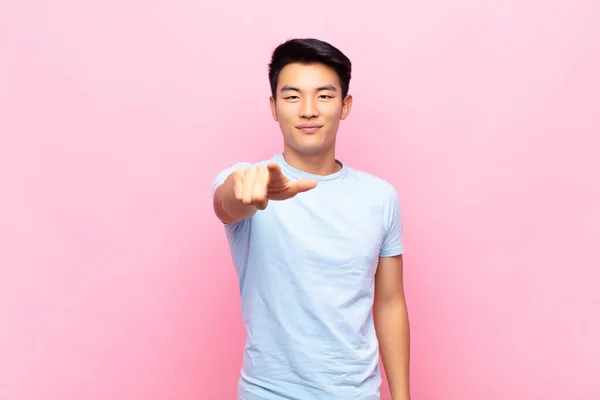 young chinese man pointing at camera with a satisfied, confident, friendly smile, choosing you against flat color wall