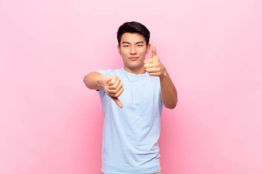 Genç Çinli bir adam kafası karışmış, bilgisiz ve kararsız hissediyor, farklı seçeneklerde iyi ve kötü şeyleri tartıyor ya da düz renkli duvarlara karşı seçenekleri tartıyor.