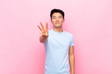 Genç Çinli adam gülümsüyor ve arkadaş canlısı görünüyor. Üç ya da üçüncüyü gösteriyor. Elleri önde, düz renkli duvara doğru sayıyor.