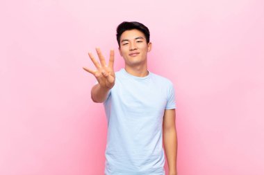 Genç Çinli adam gülümsüyor ve arkadaş canlısı görünüyor, dört ya da dört numarayı gösteriyor, düz renkli duvara doğru sayıyor.
