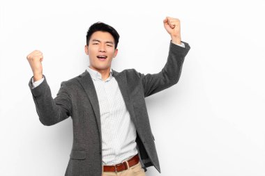 Genç Çinli adam muzaffer bir şekilde bağırıyor, heyecanlı, mutlu ve şaşırmış görünüyor, düz renkli duvara karşı kutlama yapıyor.