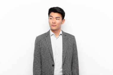 Genç Çinli adam merak ediyor, mutlu düşünceler ve fikirler düşünüyor, hayal kuruyor, düz renkli duvara karşı uzayı kopyalamaya çalışıyor.