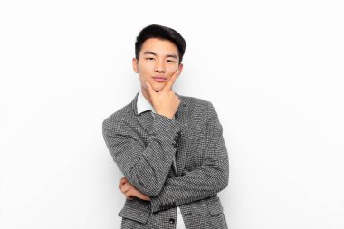 Genç Çinli bir adam ciddi, düşünceli ve güvensiz görünüyor, bir kolu çapraz ve çene üzerinde, düz renkli duvarla seçenekleri tartıyor