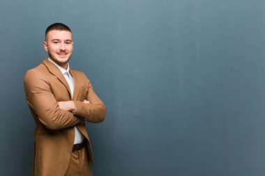 Genç yakışıklı adam kameraya çapraz kollarıyla gülümsüyor ve mutlu, kendinden emin, tatmin olmuş bir yüz ifadesi düz duvara karşı yanal bir bakış.