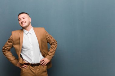 Genç yakışıklı adam mutlu, neşeli ve kendinden emin görünüyor. Gururla gülümsüyor ve kalçalarını düz arkaplana yaslıyor.