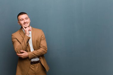 Genç yakışıklı adam mutlu bir şekilde gülümsüyor ve hayal kuruyor ya da şüphe ediyor, düz arkaplana bakıyor.