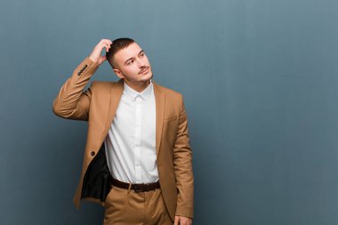 Genç yakışıklı adam şaşkın ve şaşkın hissediyor, başını kaşıyor ve düz arkaplana bakıyor.