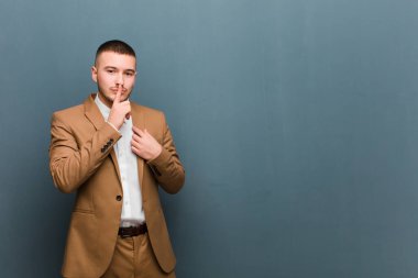 Genç ve yakışıklı adam ciddi görünüyor ve parmaklarını dudaklarına bastırıp sessizlik ya da sessizlik talep ediyor, düz arkaplan karşısında bir sır saklıyor.