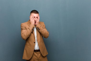 Genç yakışıklı iş adamı korkmuş ya da utanmış hissediyor, gözleri yarı kapalı ve düz arkaplan karşısında dikizliyor ya da dikizliyor.