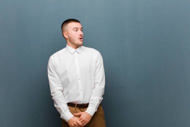 Genç yakışıklı iş adamı şok olmuş, mutlu, şaşkın ve şaşırmış hissediyor, düz arkaplana karşı ağzı açık bir şekilde yan tarafa bakıyor.