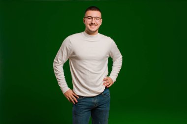Genç ve yakışıklı bir adam mutlu bir şekilde gülümsüyor, eli kalçasında ve kendinden emin, pozitif, gururlu ve dost canlısı bir tavırla düz arkaplana karşı.