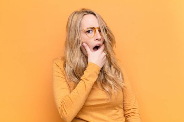 Genç ve güzel sarışın kadın ağzı ve gözleri sonuna kadar açık ve eli çenesinde, hoş olmayan bir şokta, düz renk duvara karşı ne ya da wow diyor