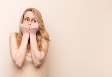 Genç, güzel, sarışın kadın endişeli, stresli ve korkmuş görünüyor. Tırnaklarını yiyor ve düz renkli duvara karşı yanal fotokopi çekmeye çalışıyor.