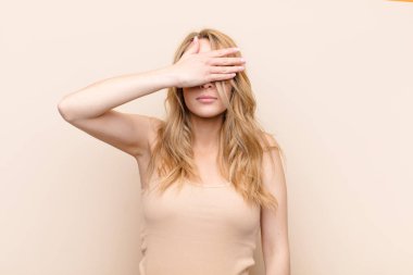 Genç, güzel, sarışın bir kadın tek eliyle gözlerini kapatıyor. Korkmuş ya da endişeli, merak ediyor ya da kör bir şekilde düz renkli duvara karşı bir sürpriz bekliyor.