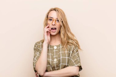 Genç ve güzel sarışın kadın şok ve inançsızlık içinde, elleri yanağında ve kolları çaprazlanmış, sersemlemiş ve düz renkli duvara karşı hayrete düşmüş.