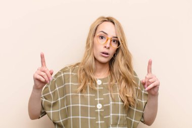 Şok olmuş, şaşkın ve ağzı açık genç, sarışın bir kadın, iki eliyle yukarıya bakıyor düz renkli duvara uzayı kopyalamak için.