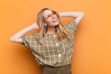 Genç ve güzel sarışın kadın gülümsüyor ve rahatlıyor, tatmin oluyor ve kaygısız, pozitif gülüyor ve düz renkli duvara karşı ürperiyor.