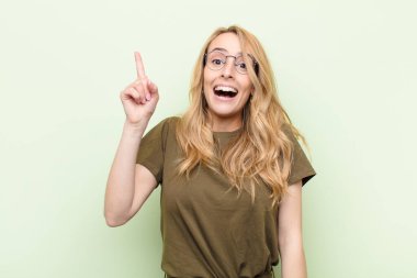Genç ve güzel sarışın kadın bir fikri fark ettikten sonra kendini mutlu ve heyecanlı bir dahi gibi hissediyor. Neşeli bir şekilde parmağını kaldırıyor. Evreka! yassı renk duvarına karşı