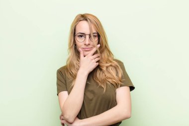 Genç, güzel sarışın kadın ciddi, düşünceli ve güvensiz görünüyor, bir kolu çapraz ve çene üzerinde, düz renkli duvarla seçenekleri tartıyor