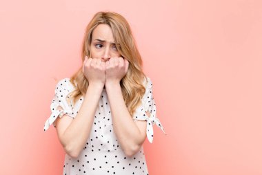 Genç, güzel, sarışın kadın endişeli, stresli ve korkmuş görünüyor. Tırnaklarını yiyor ve düz renkli duvara karşı yanal fotokopi çekmeye çalışıyor.