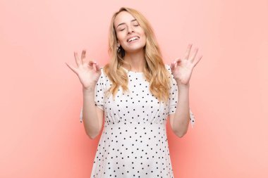 Genç ve güzel sarışın kadın konsantre olmuş ve meditasyon yapıyor, tatmin olmuş ve rahatlamış hissediyor, düşünüyor ya da düz renkli duvara karşı bir seçim yapıyor.