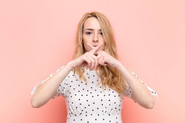 Genç ve güzel sarışın kadın ciddi görünüyor ve reddedilmek için iki parmağının önde olması hoşnutsuz, düz renkli duvara karşı sessizlik istiyor.