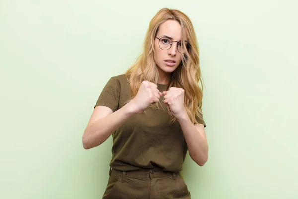 young pretty blonde woman looking confident, angry, strong and aggressive, with fists ready to fight in boxing position against flat color wall
