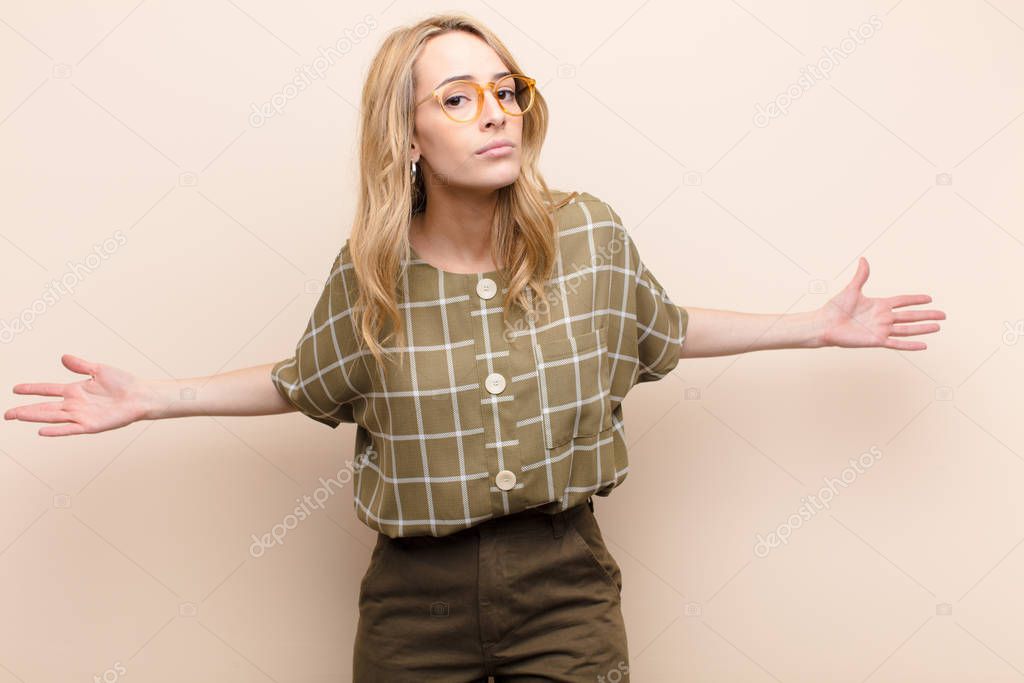 young pretty blonde woman feeling clueless and confused, having no idea, absolutely puzzled with a dumb or foolish look against flat color wall