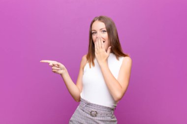 Genç kızıl saçlı kadın kendini mutlu, şaşkın ve şaşırmış hissediyor. Ağzını eliyle kapatıyor ve düz duvara karşı yanal fotokopi alanını gösteriyor.