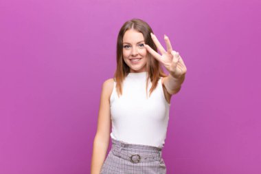 Genç kızıl saçlı kadın gülümsüyor ve arkadaş canlısı görünüyor. Üç ya da üç numarayı gösteriyor. Elleri önde, düz duvara doğru sayıyor.