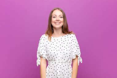 Genç kızıl saçlı kadın mutlu ve şapşal görünüyor. Geniş, eğlenceli, kaçık bir gülüşü ve düz duvarda gözleri var.