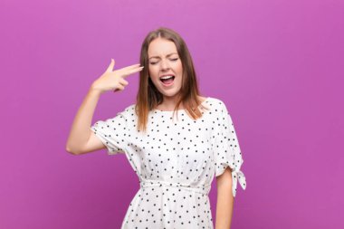 Genç kızıl saçlı kadın mutsuz ve stresli görünüyor, intihar jesti elle silah işareti yapıyor, kafasını düz duvara doğrultuyor.