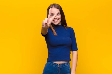 Genç kızıl saçlı kadın gülümsüyor ve arkadaş canlısı görünüyor. İki ya da iki numarayı gösteriyor. Elleri önde, düz duvara doğru sayıyor.