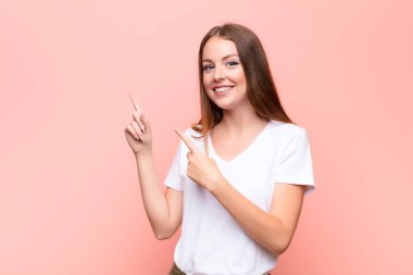 Genç kızıl saçlı kadın mutlu bir şekilde gülümsüyor ve her iki elinde de düz duvara karşı fotokopi uzayındaki nesneleri gösteriyor.