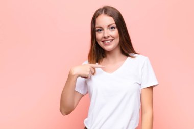 Genç kızıl saçlı kadın mutlu, gururlu ve şaşırmış görünüyor, neşeli bir şekilde kendini gösteriyor, kendinden emin ve düz duvara karşı kibirli hissediyor.