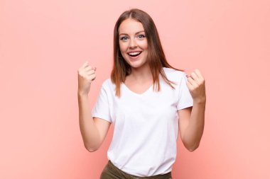 Genç kızıl saçlı kadın mutlu, şaşırmış ve gururlu hissediyor, bağırıyor ve başarıyı kutluyor düz duvara karşı büyük bir gülümsemeyle.